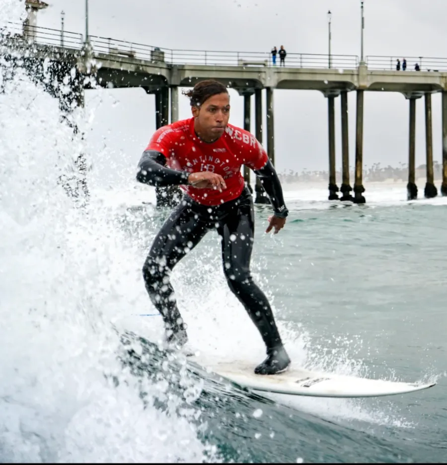 Kayiita surfing a wave beside Huntington Beach Pier in a red contest jersey.